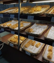 A display of freshly baked pastries is arranged in woven baskets on shelving units. The pastries have golden-brown, flaky crusts and are placed on white parchment paper. Price tags are visible, with various prices marked, indicating different types of pastries available.