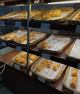 A display of freshly baked pastries is arranged in woven baskets on shelving units. The pastries have golden-brown, flaky crusts and are placed on white parchment paper. Price tags are visible, with various prices marked, indicating different types of pastries available.