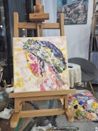An easel holds a vibrant, Impressionist-style painting depicting a side profile of a figure with a floral headdress. The background is a mix of warm tones with visible brushstrokes. The painting is surrounded by art supplies, including a palette with various colors and brushes.