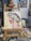 An easel holds a vibrant, Impressionist-style painting depicting a side profile of a figure with a floral headdress. The background is a mix of warm tones with visible brushstrokes. The painting is surrounded by art supplies, including a palette with various colors and brushes.