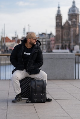 A man wearing a black jacket, white pants, and a beige cap is sitting on a suitcase outdoors. He appears to be looking to the side, and a black backpack is situated in front of him. In the background, there is a blurred view of an ornate building and other structures.