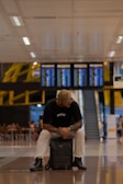 A person wearing casual attire, including a cap, black t-shirt, white pants, and boots, sits on a backpack in a modern airport terminal. Behind him are digital flight information boards displaying numerous flight details. The floor is reflective, and the ceiling has a pattern of lights. In the background, there are people seated at tables and an escalator leading to another level.