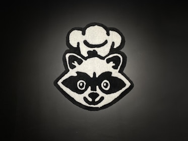 A monochrome cartoon-style graphic of a raccoon wearing a chef's hat. The image is centered against a dark background, with bold black outlines and white fill contrasting sharply against the backdrop.