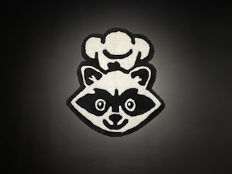 A monochrome cartoon-style graphic of a raccoon wearing a chef's hat. The image is centered against a dark background, with bold black outlines and white fill contrasting sharply against the backdrop.