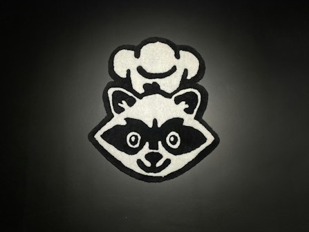 A monochrome cartoon-style graphic of a raccoon wearing a chef's hat. The image is centered against a dark background, with bold black outlines and white fill contrasting sharply against the backdrop.