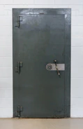 A large, heavy metal vault door set within a concrete or brick wall. The door features a circular combination lock and a metal handle bar. The surface has a slightly textured, industrial appearance.