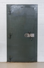 A large, heavy metal vault door set within a concrete or brick wall. The door features a circular combination lock and a metal handle bar. The surface has a slightly textured, industrial appearance.