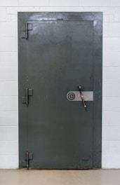 A large, heavy metal vault door set within a concrete or brick wall. The door features a circular combination lock and a metal handle bar. The surface has a slightly textured, industrial appearance.