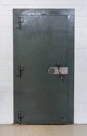 A large, heavy metal vault door set within a concrete or brick wall. The door features a circular combination lock and a metal handle bar. The surface has a slightly textured, industrial appearance.