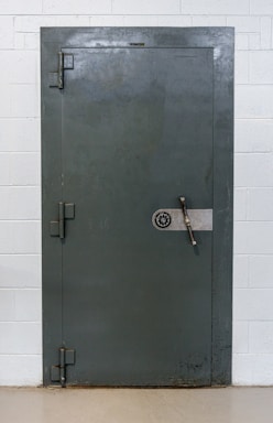 A large, heavy metal vault door set within a concrete or brick wall. The door features a circular combination lock and a metal handle bar. The surface has a slightly textured, industrial appearance.