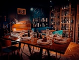 A warmly lit dining area features a table set elegantly with plates, napkins, and wine glasses. In the background, a vintage piano rests against a dark wall with a decorative arch-like window, surrounded by shelves stacked with various wine bottles. Comfortable chairs and rich wooden flooring enhance the cozy atmosphere.