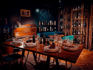 A warmly lit dining area features a table set elegantly with plates, napkins, and wine glasses. In the background, a vintage piano rests against a dark wall with a decorative arch-like window, surrounded by shelves stacked with various wine bottles. Comfortable chairs and rich wooden flooring enhance the cozy atmosphere.