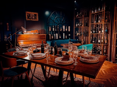 A warmly lit dining area features a table set elegantly with plates, napkins, and wine glasses. In the background, a vintage piano rests against a dark wall with a decorative arch-like window, surrounded by shelves stacked with various wine bottles. Comfortable chairs and rich wooden flooring enhance the cozy atmosphere.