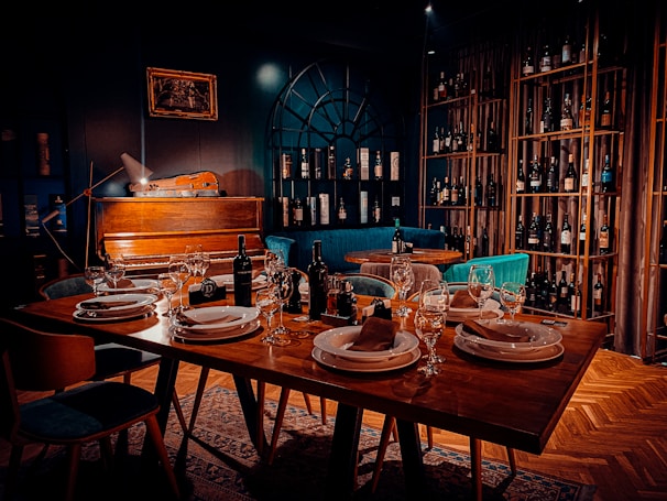 A warmly lit dining area features a table set elegantly with plates, napkins, and wine glasses. In the background, a vintage piano rests against a dark wall with a decorative arch-like window, surrounded by shelves stacked with various wine bottles. Comfortable chairs and rich wooden flooring enhance the cozy atmosphere.