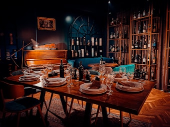 A warmly lit dining area features a table set elegantly with plates, napkins, and wine glasses. In the background, a vintage piano rests against a dark wall with a decorative arch-like window, surrounded by shelves stacked with various wine bottles. Comfortable chairs and rich wooden flooring enhance the cozy atmosphere.