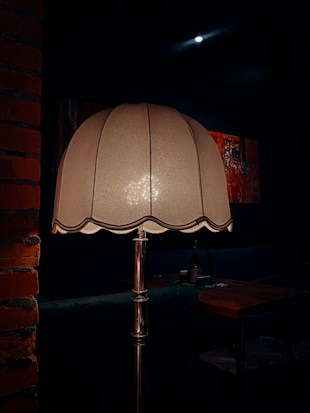 A warmly lit, vintage-style lamp stands prominently in a dimly lit room. The lamp has a fabric shade with scalloped edges and is supported by a metallic stand. In the background, a wooden table with a bottle and glasses can be seen, alongside some abstract artwork on the dark walls. The ambiance is cozy and intimate.