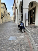 A narrow cobblestone street is lined with buildings featuring arched doorways and traditional architecture. A blue scooter is parked to the side, adding a modern touch to the historic scene. The street is empty, conveying a peaceful and quiet atmosphere.