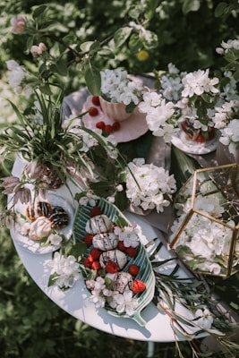 An elegantly arranged garden table features a display of white blossoms, fresh raspberries, and delectable desserts on a long green plate. The table is surrounded by green foliage, enhancing the natural, outdoor setting. Some elements like a pink tea cup and a glass terrarium add to the sophisticated yet rustic decor.