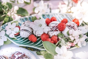 A decorative plate with white desserts topped with chocolate drizzle and raspberries. Surrounded by white flowers and green leaves, creating a fresh, elegant presentation.
