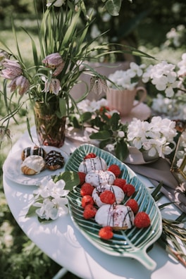A decorative table setup featuring white and black desserts with raspberries arranged on a green leaf-shaped platter. Beside it are pastries topped with icing and chocolate. In the background, a vase holds tall grasses and checkerboard-patterned flowers. Surrounding the scene are white blossoms, contributing to an elegant and natural aesthetic.
