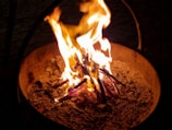 A fire burning in a metal container with bright yellow and orange flames rising from a bed of ashes and partially burnt wood.