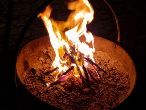A fire burning in a metal container with bright yellow and orange flames rising from a bed of ashes and partially burnt wood.