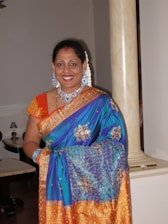 A woman stands indoors wearing a colorful sari adorned with intricate patterns, along with matching jewelry including a necklace, earrings, and bangles. The sari features vibrant blue and orange hues with gold embroidery. Her hair is styled with decorations, and she is smiling warmly.
