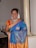 A woman stands indoors wearing a colorful sari adorned with intricate patterns, along with matching jewelry including a necklace, earrings, and bangles. The sari features vibrant blue and orange hues with gold embroidery. Her hair is styled with decorations, and she is smiling warmly.