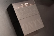 A black box with an elegant, embossed design is prominently featured. The top part of the box displays the word 'ECLIPSE' in capital letters followed by a description of flavors including dark chocolate, burnt sugar, and smoked vanilla bean. The lower portion shows the words 'ONYX COFFEE' with a sleek, modern typography.