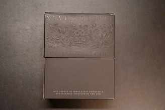 A dark-colored box with embossed text and floral designs. It includes the phrases 'Never Settle For Good Enough' and 'Our coffee is impeccably sourced & sustainably roasted by the sun.' The overall design is simple and elegant.