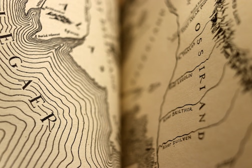 A detailed close-up of a sepia-toned map featuring contour lines and legible text, including geographic names such as Ossiriand and River Brilthor. The focus transitions smoothly from sharp details to a softer blur, emphasizing the intricacy of the cartographic design.