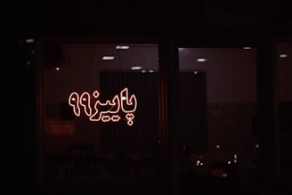 A dimly lit room seen through a window, featuring illuminated Persian script that reads 'پاییز 99'. The ambient lighting creates a warm glow, and some furniture or objects are vaguely visible inside.