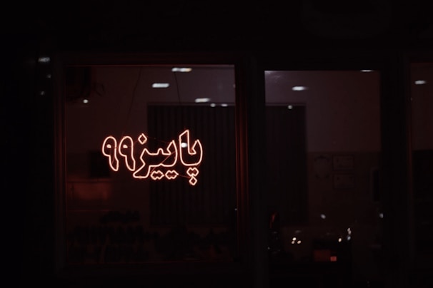 A dimly lit room seen through a window, featuring illuminated Persian script that reads 'پاییز 99'. The ambient lighting creates a warm glow, and some furniture or objects are vaguely visible inside.