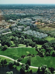 Aerial view of a cityscape blending urban and natural environments, featuring numerous residential buildings interspersed with lush green parks. The foreground includes well-maintained grassy areas and walking paths, leading to a densely built urban area in the background.