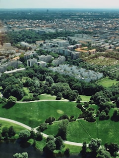Aerial view of a cityscape blending urban and natural environments, featuring numerous residential buildings interspersed with lush green parks. The foreground includes well-maintained grassy areas and walking paths, leading to a densely built urban area in the background.