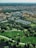 Aerial view of a cityscape blending urban and natural environments, featuring numerous residential buildings interspersed with lush green parks. The foreground includes well-maintained grassy areas and walking paths, leading to a densely built urban area in the background.