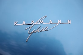 The image features the logo and emblem 'Karmann Ghia' on a light blue background. The logo is written in metallic silver with an elegant, stylized font that stands out against the contrasting background.