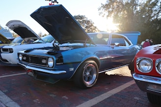 A classic blue muscle car with its hood open is parked on a brick surface, surrounded by other vintage vehicles. The sun shines in the background, casting a warm glow and creating lens flares. Trees and event attendees are visible in the background.