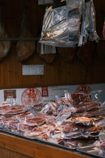 Various cuts of meat are vacuum-sealed in clear plastic packaging, displayed on a wooden counter. Above the counter, additional packaged fish and meats hang. The background consists of a wooden wall with decorative elements and muted lighting.