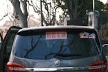 A close-up view of the rear window of a dark-colored car, displaying a sign with Chinese characters. The window reflects bare tree branches, indicating a winter setting. There is also a small sticker featuring cartoon figures on the window.