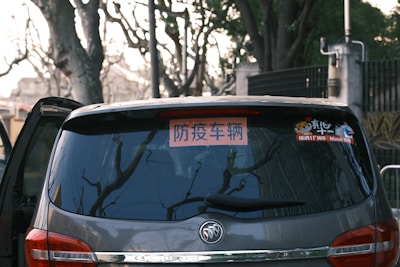 A close-up view of the rear window of a dark-colored car, displaying a sign with Chinese characters. The window reflects bare tree branches, indicating a winter setting. There is also a small sticker featuring cartoon figures on the window.