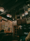 A cozy indoor space with wooden elements and warm lighting. The ceiling features multiple hanging lamps, and there is a section of the wall covered in colorful sticky notes and photos. An air conditioning unit is mounted on the wall. There is a wooden door with glass panels and some shelving with plants and decorative items. There are people sitting at a table, partially visible in the bottom corners of the image.