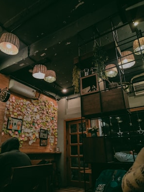 A cozy indoor space with wooden elements and warm lighting. The ceiling features multiple hanging lamps, and there is a section of the wall covered in colorful sticky notes and photos. An air conditioning unit is mounted on the wall. There is a wooden door with glass panels and some shelving with plants and decorative items. There are people sitting at a table, partially visible in the bottom corners of the image.