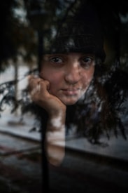 A person gazes through a window, their face partially obscured by reflections of trees and the outside environment. The expression is contemplative, with one hand resting on their chin. The scene is a blend of indoors and outdoors, creating an introspective atmosphere.