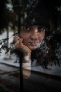 A person gazes through a window, their face partially obscured by reflections of trees and the outside environment. The expression is contemplative, with one hand resting on their chin. The scene is a blend of indoors and outdoors, creating an introspective atmosphere.