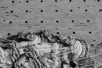 A stone relief sculpture depicting a dynamic scene of horses in motion. The artwork exhibits detailed craftsmanship, capturing the musculature and movement of the horses. This classical style is set against a backdrop of uniform, horizontal brickwork with evenly spaced, small square openings.