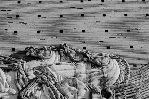 A stone relief sculpture depicting a dynamic scene of horses in motion. The artwork exhibits detailed craftsmanship, capturing the musculature and movement of the horses. This classical style is set against a backdrop of uniform, horizontal brickwork with evenly spaced, small square openings.