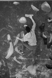 A person is engaged in indoor rock climbing, reaching up and gripping holds on a climbing wall. The setup includes various shapes and sizes of climbing holds attached to the wall. The person is wearing casual climbing attire, and the setting appears to be in a gym environment.