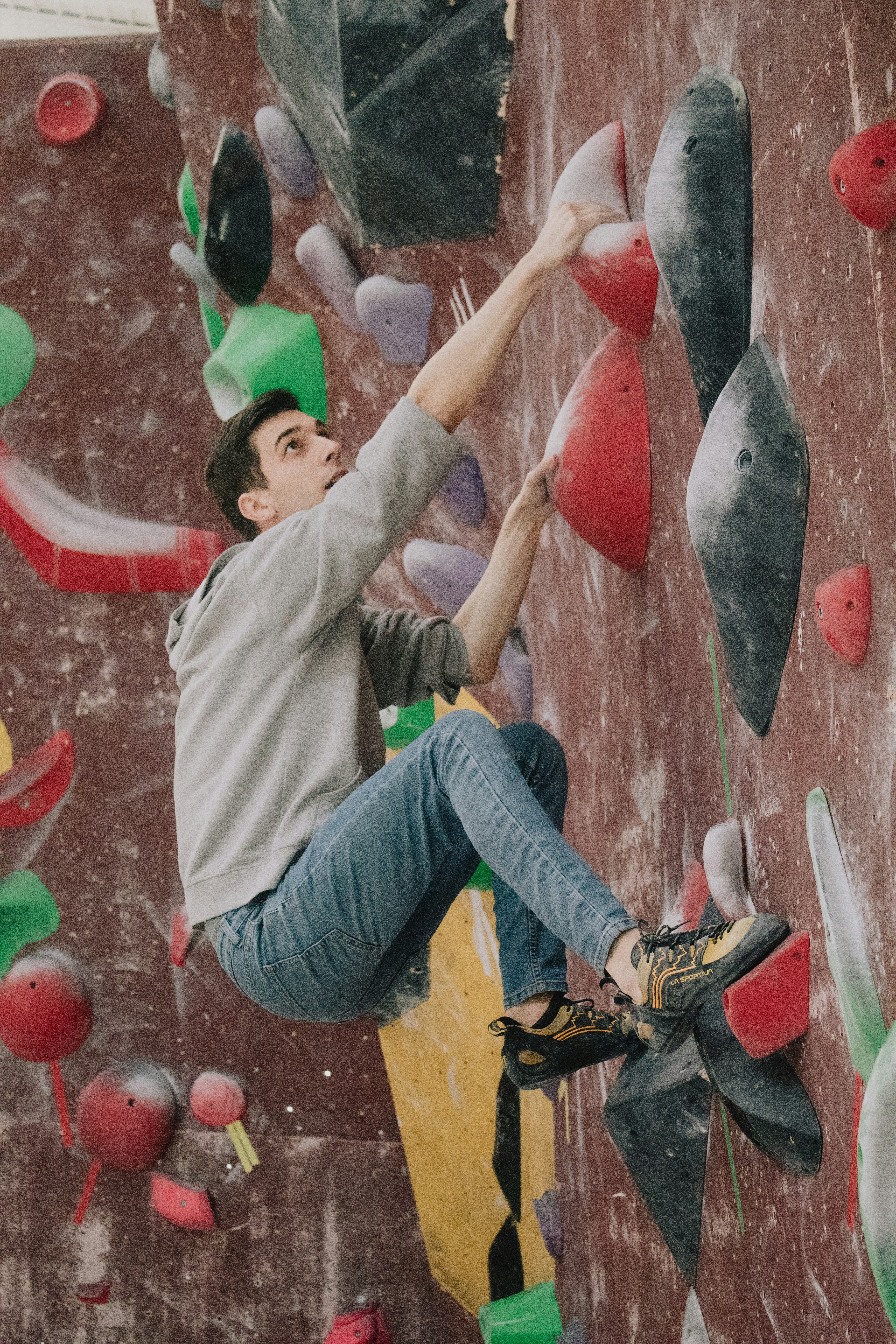 A person is engaged in indoor rock climbing on a wall covered with colorful climbing holds. The climber is wearing a gray hoodie, blue jeans, and climbing shoes, appearing focused and determined.