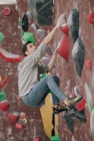A person is engaged in indoor rock climbing on a wall covered with colorful climbing holds. The climber is wearing a gray hoodie, blue jeans, and climbing shoes, appearing focused and determined.